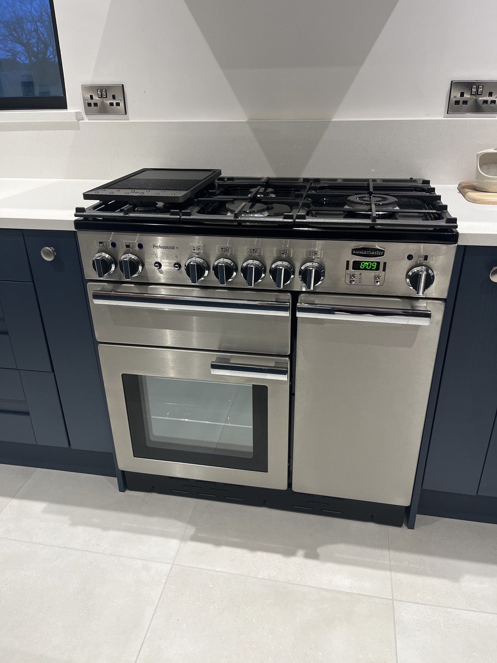 A stainless steel range cooker with a gas hob, set between navy kitchen units under a white worktop.