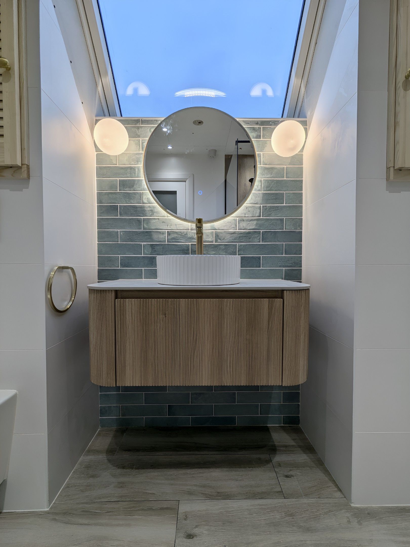 A wall-hung oak vanity with a white fluted basin and brass tap, under a round backlit mirror, with green-grey tiles and a roof window above.