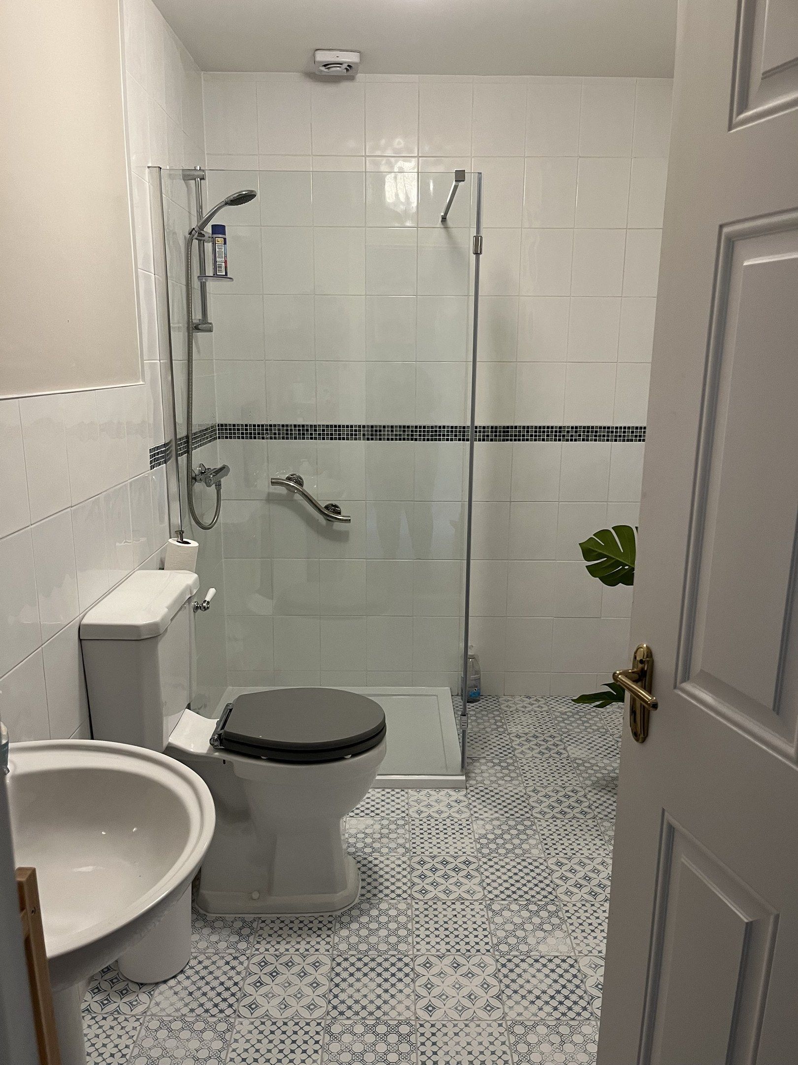 A walk-in shower with a glass panel and a grab rail, white tiles with a mosaic band, and a patterned grey and white floor.