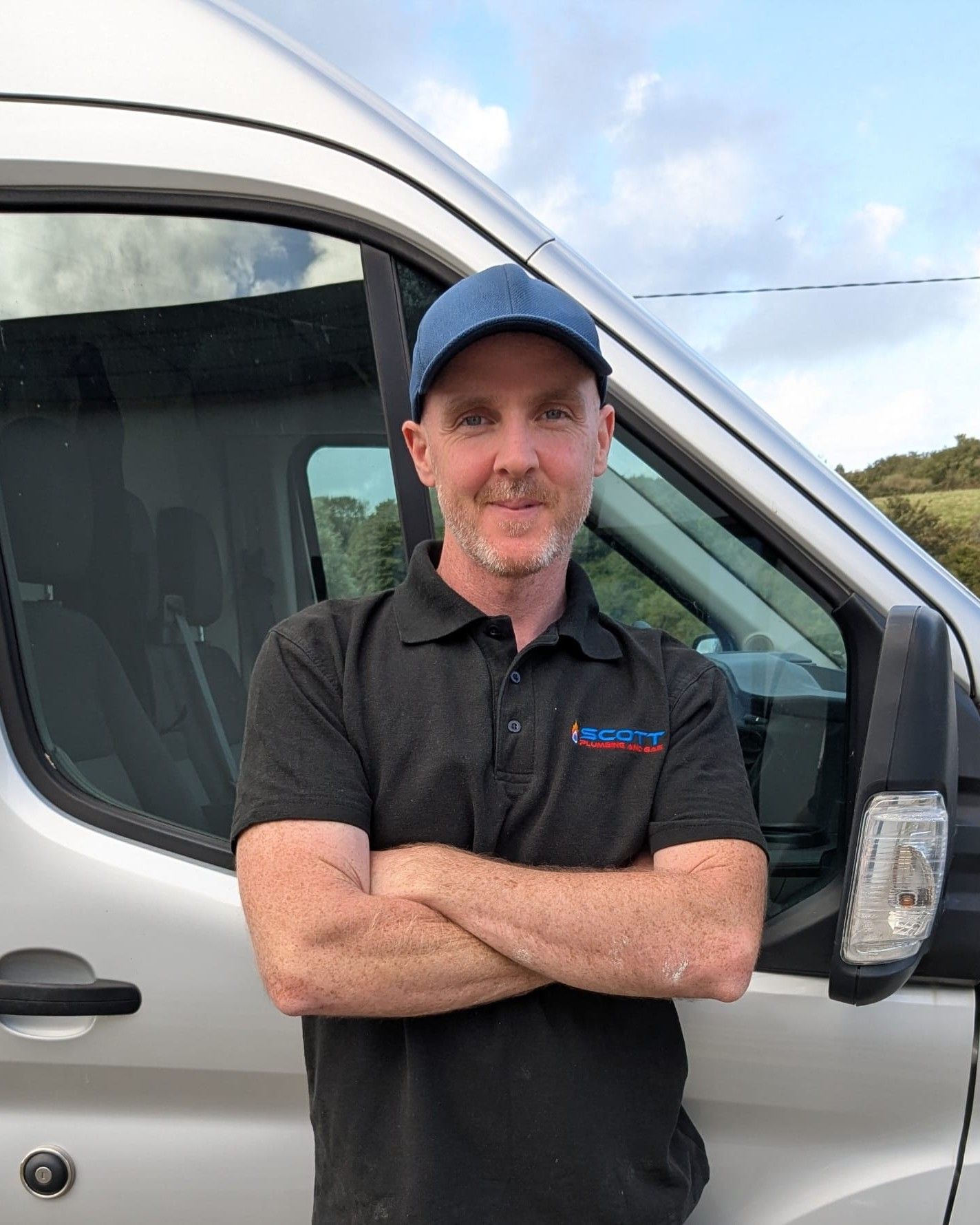 Scott Murray in a blue cap and a black work polo, arms folded, standing beside his silver van.